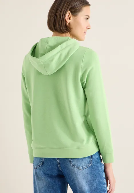 Rückansicht einer Person in grüner Kapuzenjacke und Jeans vor neutralem Hintergrund